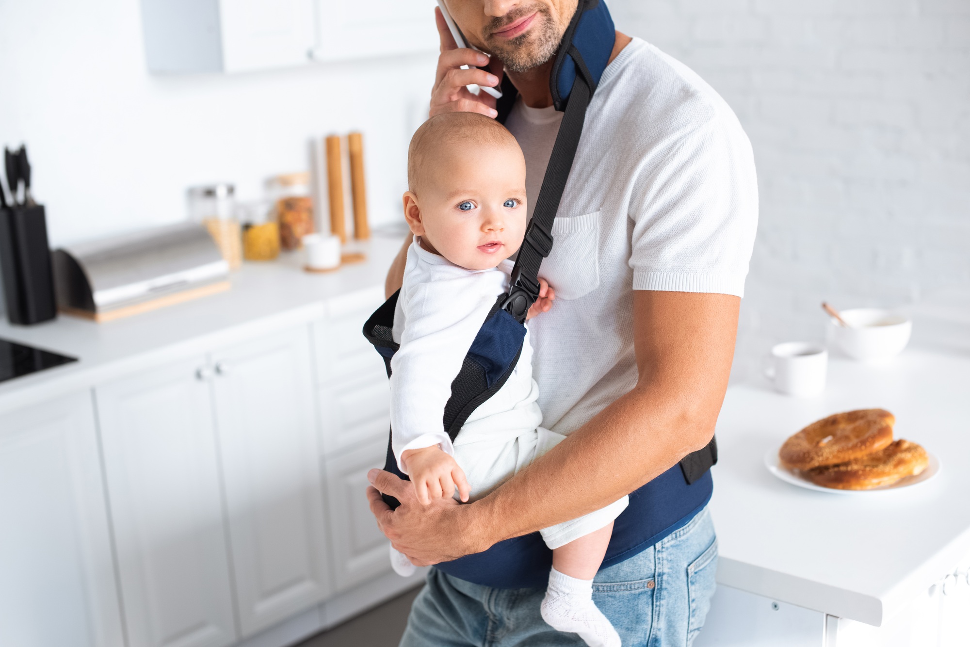 cropped view of father holding infant daughter in baby carrier and talking on smartphone